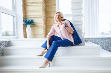 Mavi rahat takım ve pembe gömlek ev iç pencerede yakınındaki sütuna göre beyaz merdivenlerde oturan güzellik genç sarışın bir kadın kıvırcık saçlı portresi