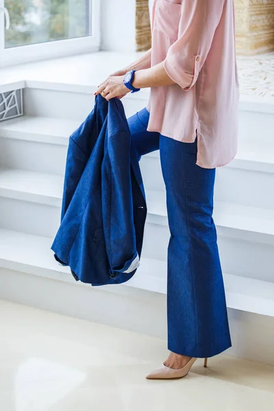 Young woman wearing blue casual suit, pink shirt and high heels standing by white stairs near window at home interior