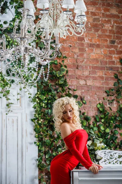 Fashion studio portrait of beautiful blond woman with curly hair and stylish makeup, wearing red evening dress, standing near white piano. Room interior with green plants on background
