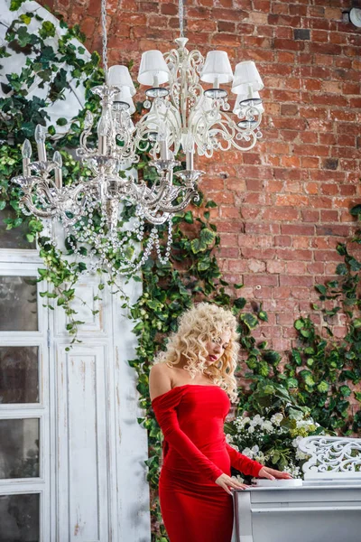 时尚工作室肖像美丽的金发女郎与卷曲的头发和时尚的妆 身穿红色晚礼服 站在附近的白色钢琴 室内与绿色植物背景 — 图库照片