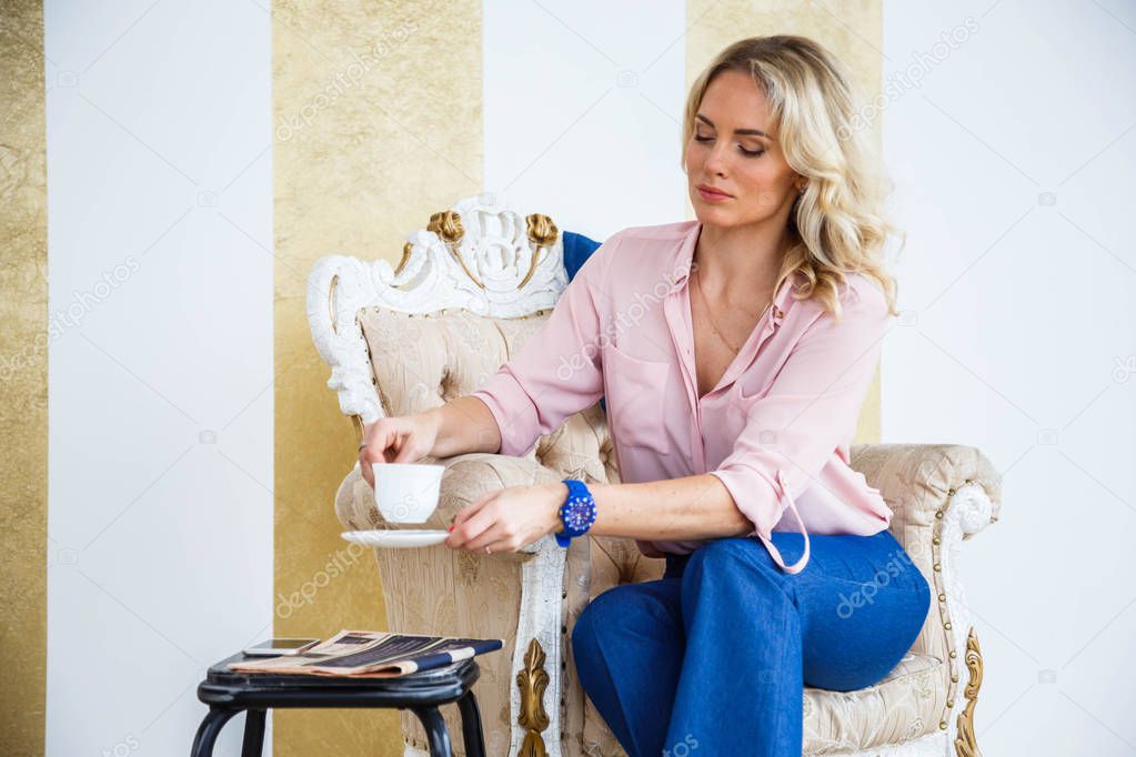 Portrait of beauty young blond woman with curly hair, wearing blue casual suit and pink shirt sitting in vintage armchair and drinking morning coffee. White and gold wall and floor at home interior background
