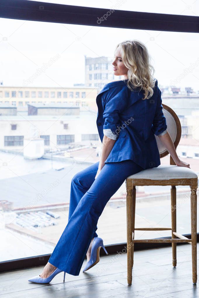 Studio portrait of beautiful blond woman with curly hair and stylish makeup, wearing blue casual suit and blue shoes on high hills, sitting on white vintage chair, blurred city view in window on background 