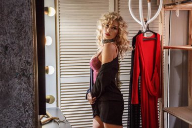 Ampulleri gardırop, ahşap kapılar ve arka plan üzerinde kırmızı elbise tarafından ışıklı ayna yakınındaki güzellik mor iç çamaşırı giyen güzel genç kadın kıvırcık sarı saçlı ayakta. 