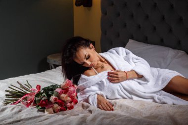 Güzellik profesyonel makyaj ile genç esmer kadın beyaz bornoz stüdyo portre. Pembe gül buketi, gri başlık arka plan ile yatakta yatarken güzel kadın