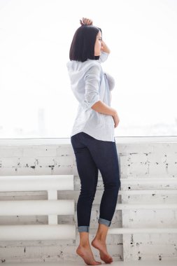 Stüdyo Daire iç pencerede yakınındaki koyu kot pantolon ve beyaz gömlek ayakta giyen güzel esmer kadın görüntüsünü