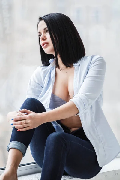 工作室拍摄的美丽的黑发女子身穿深色牛仔裤和白色衬衫坐在窗台附近的公寓室内 — 图库照片