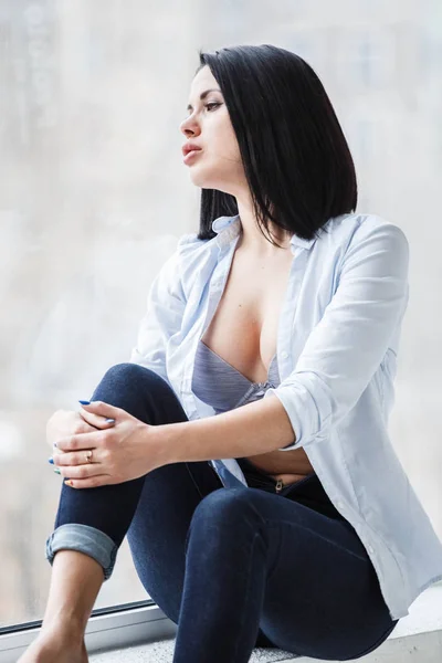 濃いジーンズとアパート室内で窓際の窓辺に座っている白いシャツを着て美しいブルネットの女性のスタジオ撮影 — ストック写真