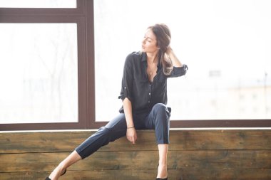 Ahşap zemin, ışıklı windows arka plan üzerinde oturan siyah rahat kıyafetler giymiş güzel genç kadın stüdyo çekim