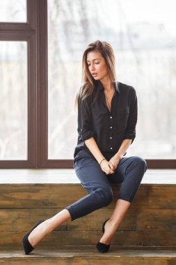 Ahşap zemin, ışıklı windows arka plan üzerinde oturan siyah rahat kıyafetler giymiş güzel genç kadın stüdyo çekim