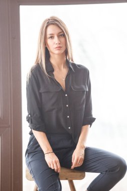 Ahşap sandalye, ışıklı pencere arka plan üzerinde oturan siyah rahat kıyafetler giymiş güzel genç kadın stüdyo çekim