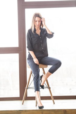 Ahşap sandalye, ışıklı pencere arka plan üzerinde oturan siyah rahat kıyafetler giymiş güzel genç kadın stüdyo çekim