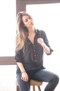 Ahşap sandalye, ışıklı pencere arka plan üzerinde oturan siyah rahat kıyafetler giymiş güzel genç kadın stüdyo çekim