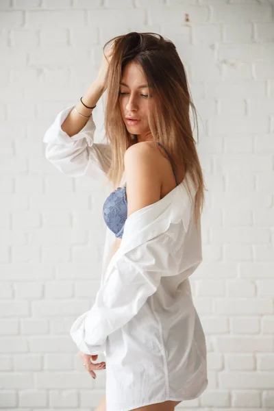 青いランジェリーと白いシャツを着て美容女性のスタジオ撮影 れんが造りの白い壁を背景に立っている若い女性 — ストック写真