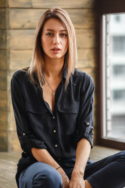 Ahşap zemin, ışıklı windows arka plan üzerinde oturan siyah rahat kıyafetler giymiş güzel genç kadın stüdyo çekim