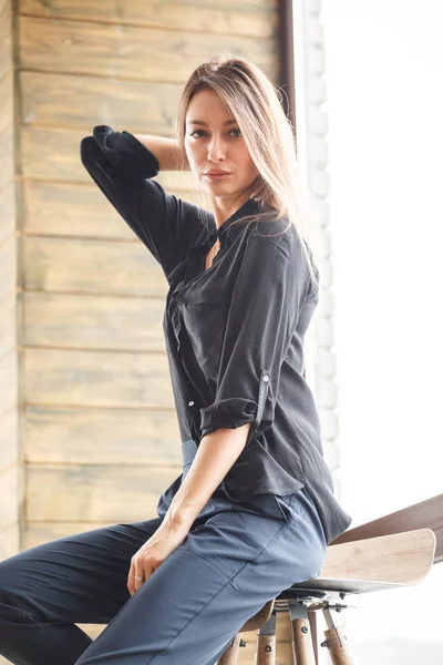 Ahşap sandalye, ışıklı pencere arka plan üzerinde oturan siyah rahat kıyafetler giymiş güzel genç kadın stüdyo çekim