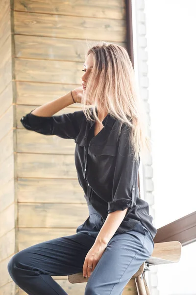 Ahşap sandalye, ışıklı pencere arka plan üzerinde oturan siyah rahat kıyafetler giymiş güzel genç kadın stüdyo çekim