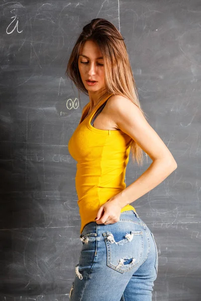 黄色の シャツとブルー ジーンズ黒板背景に立っている身に着けている美しい若い女性 — ストック写真