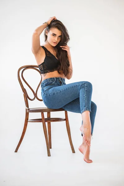 ブルー ジーンズと木製の椅子 壁の白い背景の上に座って性的な黒い下着を身に着けている美しいカーリー ブルネットの少女のスタジオ ポートレート — ストック写真