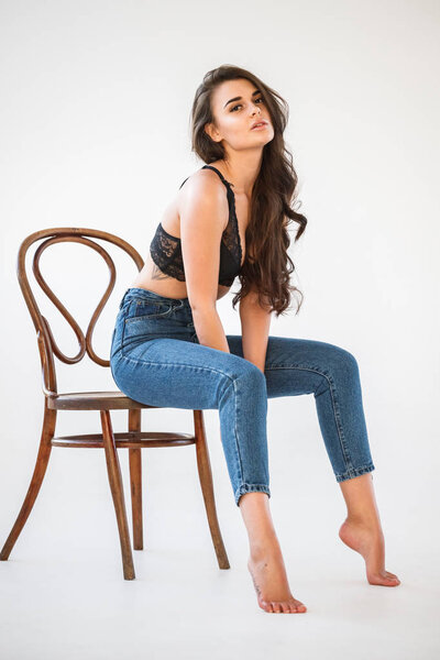 Studio portrait of beautiful curly brunette girl wearing blue jeans and sexual black underwear sitting on wooden chair, white wall background