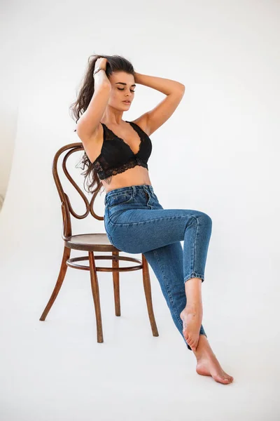 ブルー ジーンズと木製の椅子 壁の白い背景の上に座って性的な黒い下着を身に着けている美しいカーリー ブルネットの少女のスタジオ ポートレート — ストック写真