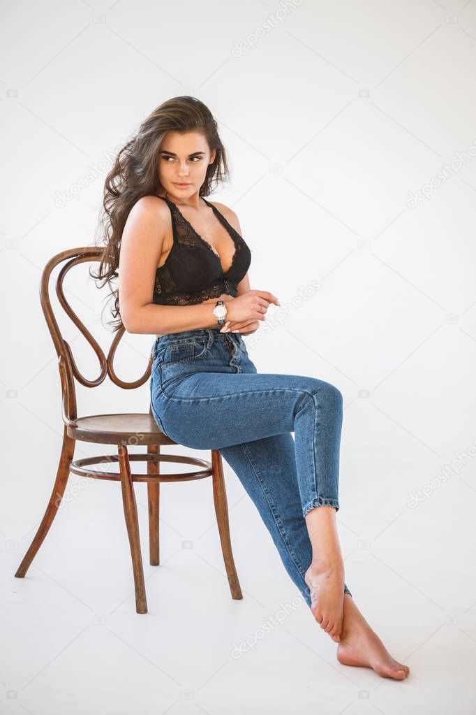 Studio portrait of beautiful curly brunette girl wearing blue jeans and sexual black underwear sitting on wooden chair, white wall background