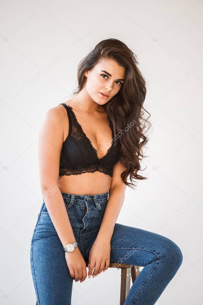 Studio portrait of beautiful curly brunette girl wearing blue jeans and sexual black underwear leaning on wooden staircase, white wall background