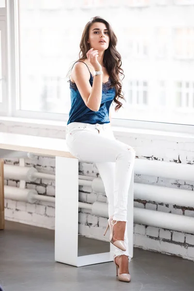年轻性感黑发女孩的肖像在蓝色内衣和白色裤子坐在桌子附近的窗口在工作室 — 图库照片
