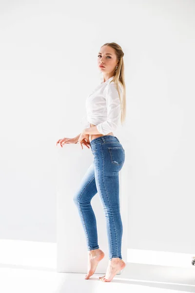 ブルー ジーンズ 白いスタジオの背景の上に立って白いワイシャツを身に着けているセクシーなブロンドの女の子の肖像画 — ストック写真