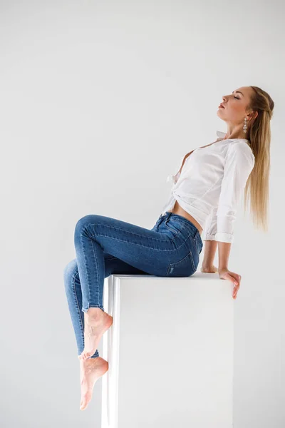 性感金发女郎的肖像穿着蓝色牛仔裤和白色衬衫坐在白色工作室背景 — 图库照片