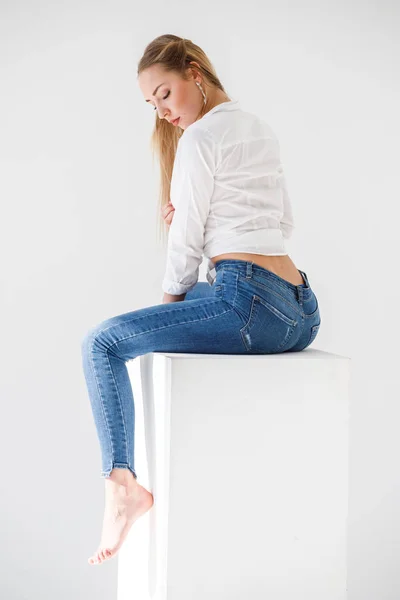 ブルー ジーンズ 白いスタジオの背景の上に座って白いワイシャツを身に着けているセクシーなブロンドの女の子の肖像画 — ストック写真