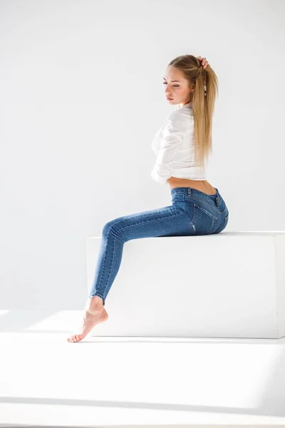 性感金发女郎的肖像穿着蓝色牛仔裤和白色衬衫坐在白色工作室背景 — 图库照片