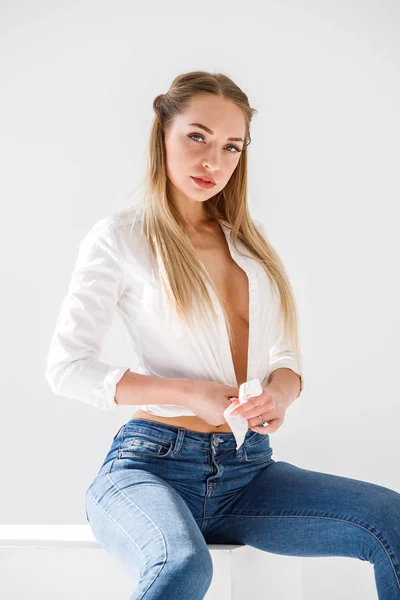 ブルー ジーンズ 白いスタジオの背景の上に座って白いワイシャツを身に着けているセクシーなブロンドの女の子の肖像画 — ストック写真