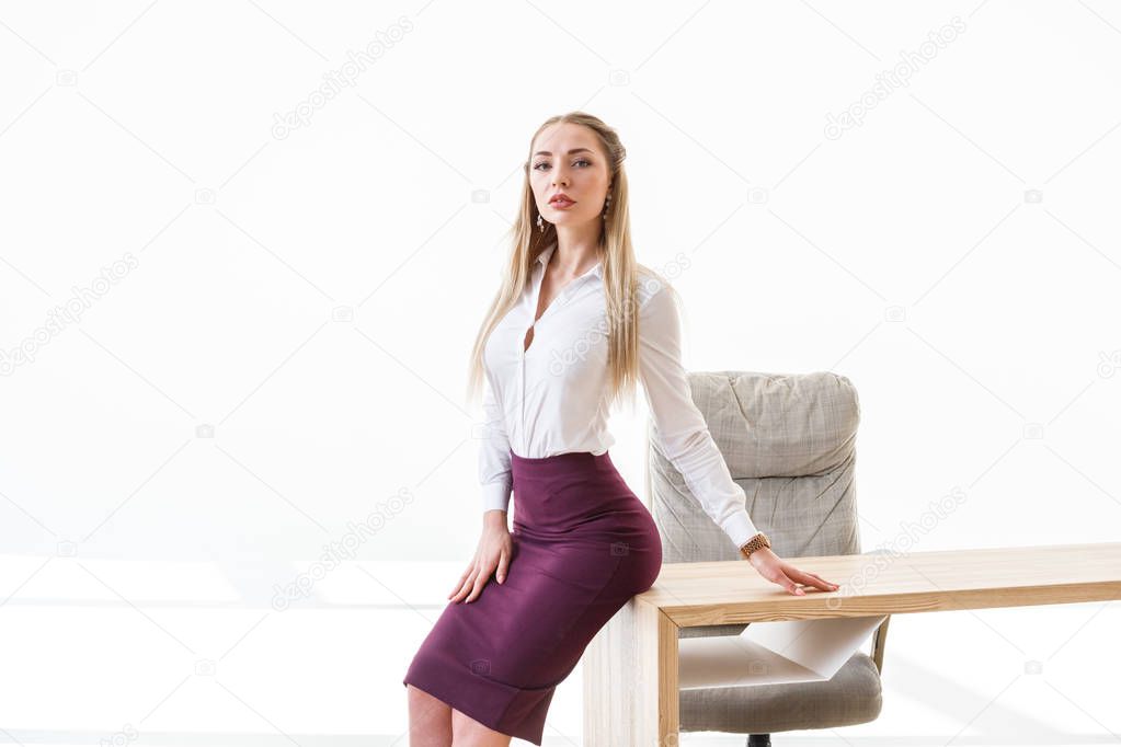 Portrait of sexy blonde girl wearing purple skirt and white shirt sitting on wooden table at modern office interior 