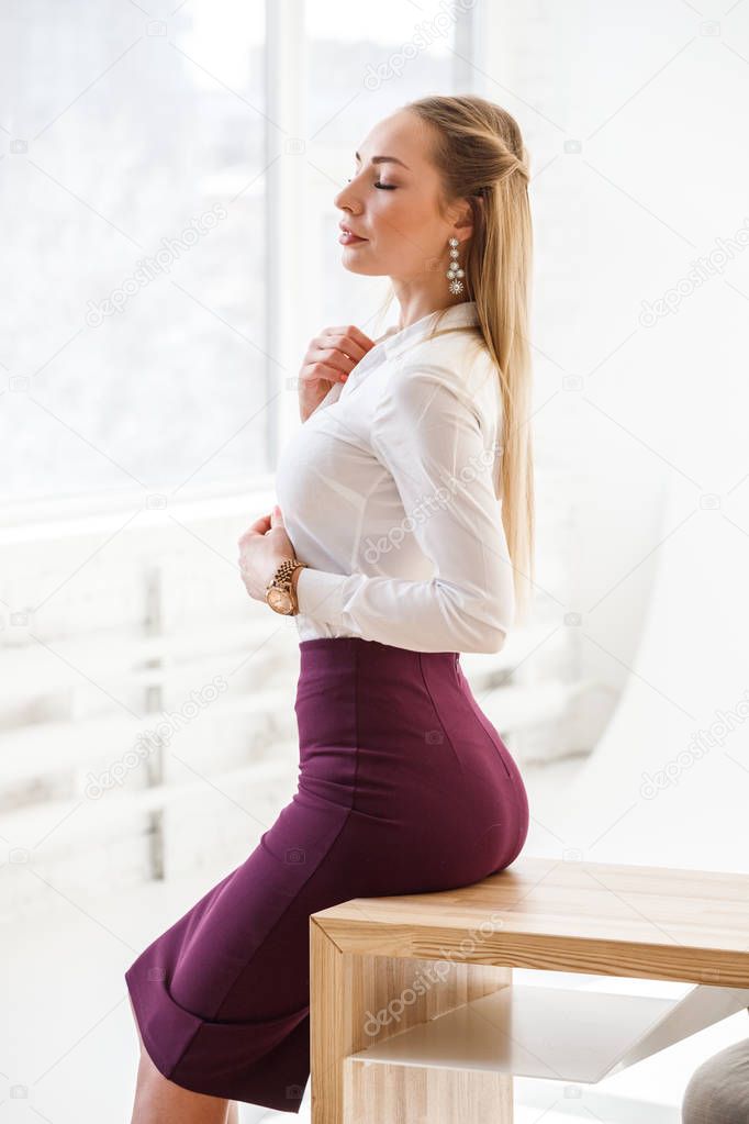 Portrait of sexy blonde girl wearing purple skirt and white shirt sitting on wooden table at modern office interior 