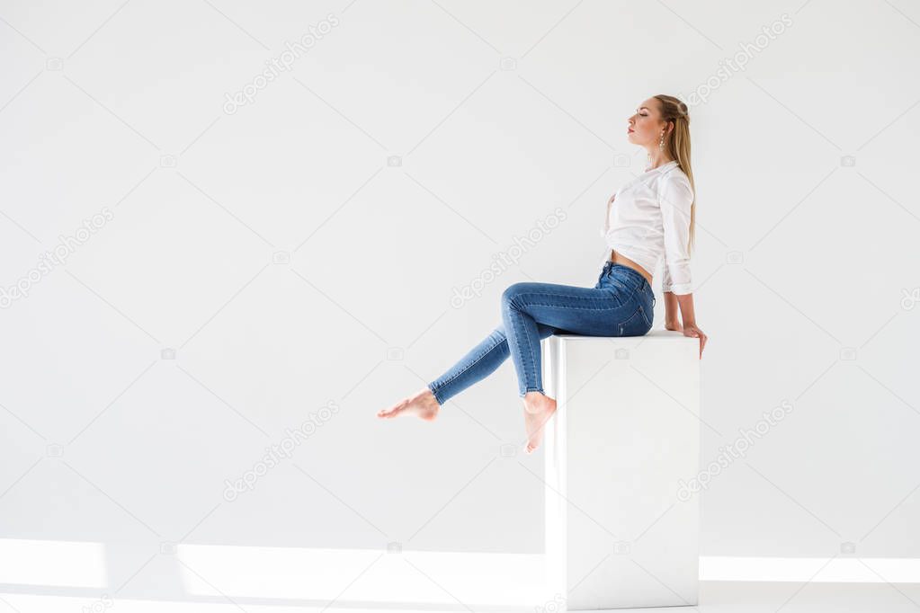 Portrait of sexy blonde girl wearing blue jeans and white shirt sitting on white studio background