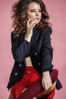Moda stüdyo kırmızı etek, siyah ceket ve tahta sandalyede oturan pembe arka plan üzerinde siyah iç çamaşırı giyen güzel kıvırcık kadın portresi