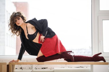 Gün ışığında ahşap pencere yatan kırmızı etek, siyah ceket ve siyah iç çamaşırı giyen güzel kıvırcık kadın moda studio portresi