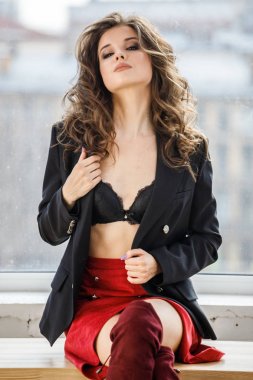 Gün ışığında ahşap pencere yatan kırmızı etek, siyah ceket ve siyah iç çamaşırı giyen güzel kıvırcık kadın moda studio portresi