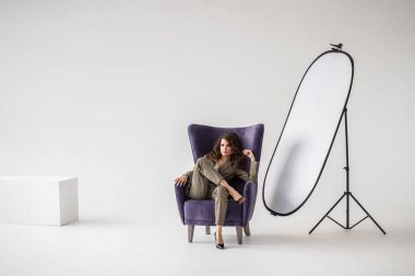 Kırmızı kemer, mor koltuk, beyaz duvar arka plan oturan sıradan takım elbise giyen güzel kıvırcık kadın moda stüdyo portre kareli 