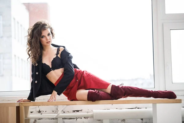 日光の下で木製の窓辺に横たわっている赤いスカート 黒のジャケットと黒のランジェリーを身に着けている美しい巻き毛女性のファッション スタジオ ポートレート — ストック写真