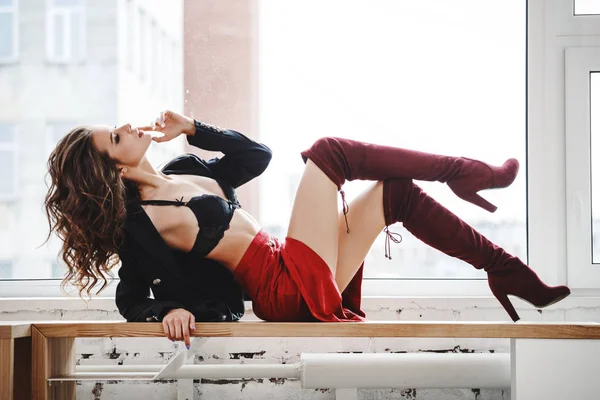 Gün ışığında ahşap pencere yatan kırmızı etek, siyah ceket ve siyah iç çamaşırı giyen güzel kıvırcık kadın moda studio portresi