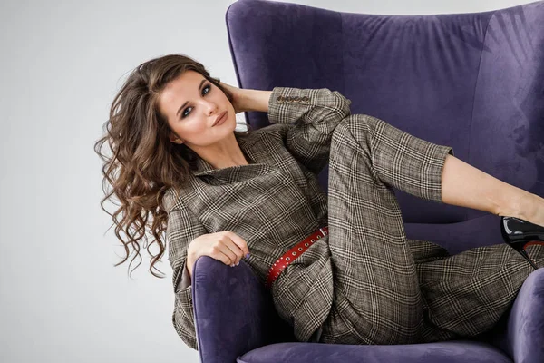 Kırmızı kemer, mor koltuk, beyaz duvar arka plan oturan sıradan takım elbise giyen güzel kıvırcık kadın moda stüdyo portre kareli 