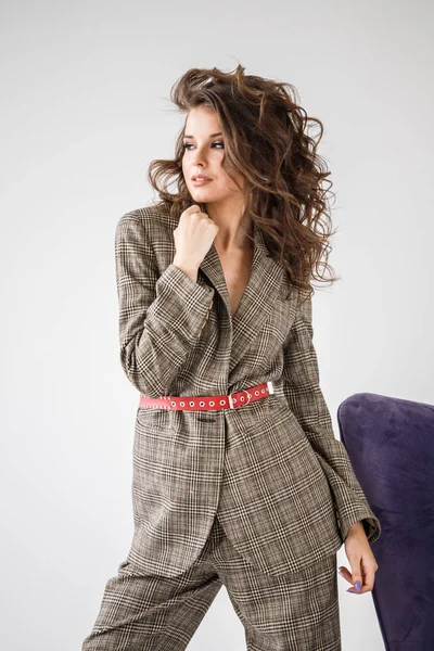 Kırmızı kemer, mor koltuk, beyaz duvar arka plan eğilerek ile rahat takım elbise giyen güzel kıvırcık kadın moda stüdyo portre kareli 