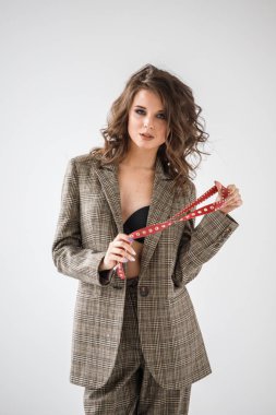 Damalı rahat takım ve siyah sütyen, ellerinde kırmızı kemer ile gri duvar arka plan üzerinde duran güzel kıvırcık kadın moda studio portresi