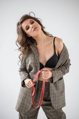 Damalı rahat takım ve siyah sütyen, ellerinde kırmızı kemer ile gri duvar arka plan üzerinde duran güzel kıvırcık kadın moda studio portresi