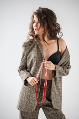 Damalı rahat takım ve siyah sütyen, ellerinde kırmızı kemer ile gri duvar arka plan üzerinde duran güzel kıvırcık kadın moda studio portresi