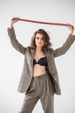Damalı rahat takım ve siyah sütyen, ellerinde kırmızı kemer ile gri duvar arka plan üzerinde duran güzel kıvırcık kadın moda studio portresi
