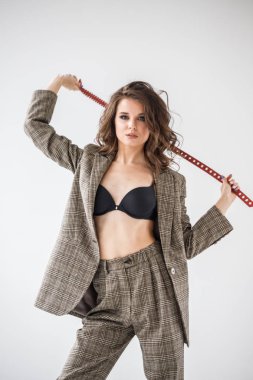 Damalı rahat takım ve siyah sütyen, ellerinde kırmızı kemer ile gri duvar arka plan üzerinde duran güzel kıvırcık kadın moda studio portresi