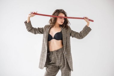 Damalı rahat takım ve siyah sütyen, ellerinde kırmızı kemer ile gri duvar arka plan üzerinde duran güzel kıvırcık kadın moda studio portresi