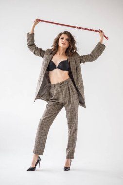 Damalı rahat takım ve siyah sütyen, ellerinde kırmızı kemer ile gri duvar arka plan üzerinde duran güzel kıvırcık kadın moda studio portresi